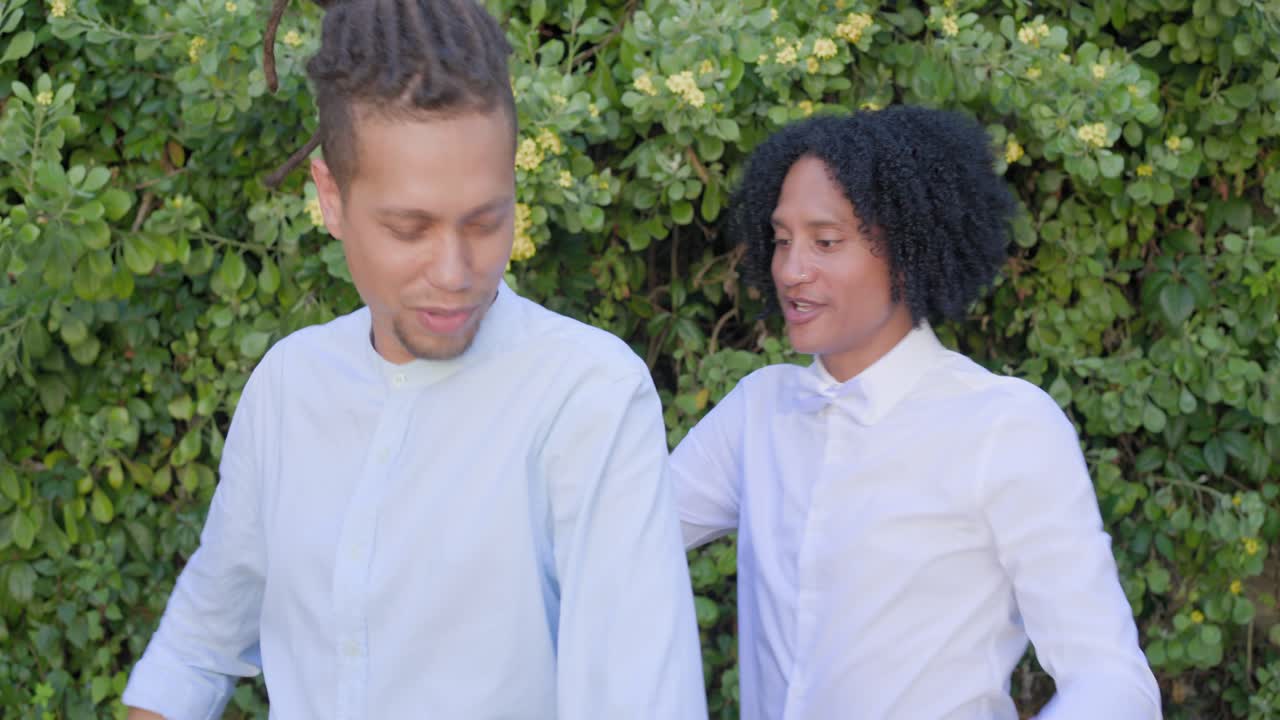 Amigos afro-americanos de meia-idade rindo, com o homem da esquerda ajustando a gravata borboleta ao lado de uma cerca viva.