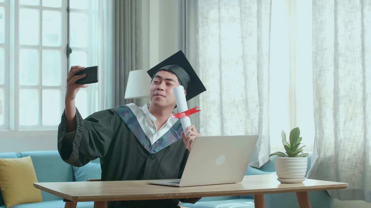 Young Asian Man Taking A Selfie While Wearing A Graduation Gown And Cap, Hand Holding University Certificate In Living Room