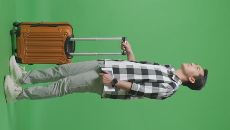 full body side view of asian male traveler with luggage holding passport and looking around while standing in the green screen background studio