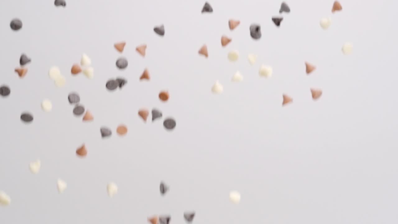 화이트 초콜릿, 버터스카치 캐러멜, 세미 스위트 초콜릿 칩이 흰색 배경에서 슬로우 모션으로 비가 내리고 있습니다.