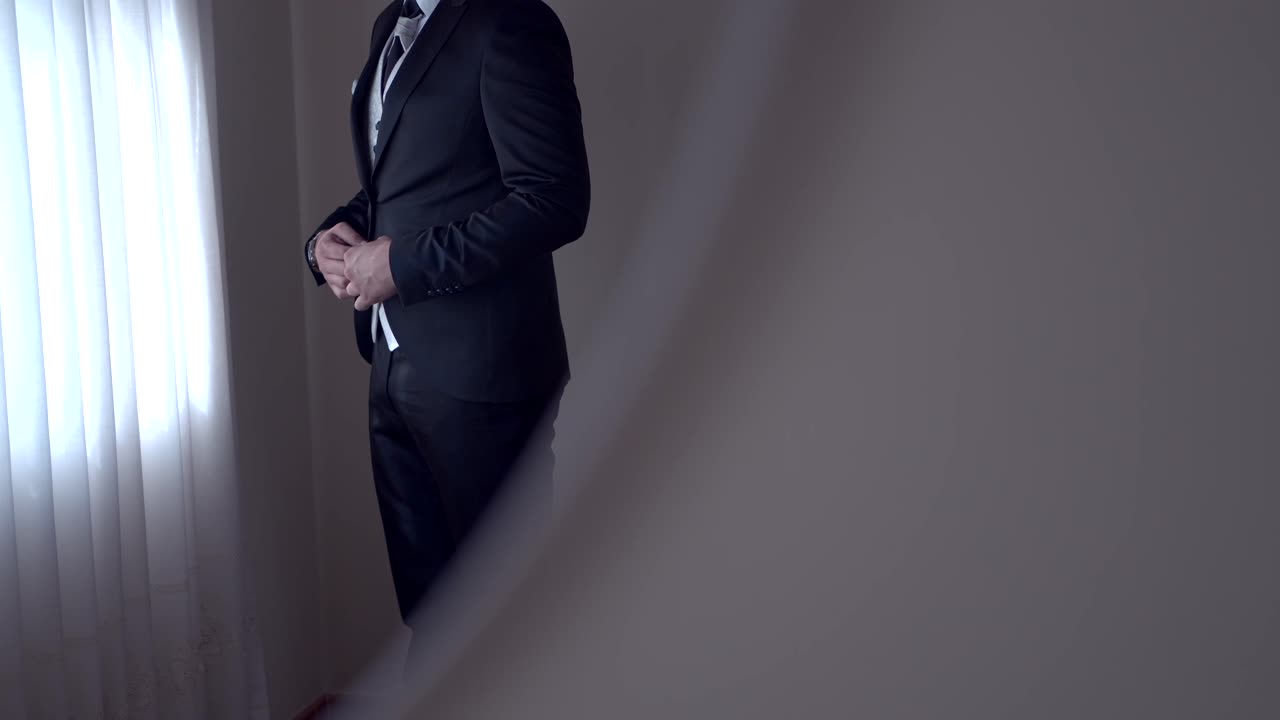 Elegant groom in a dark suit adjusting his jacket near a softly lit window