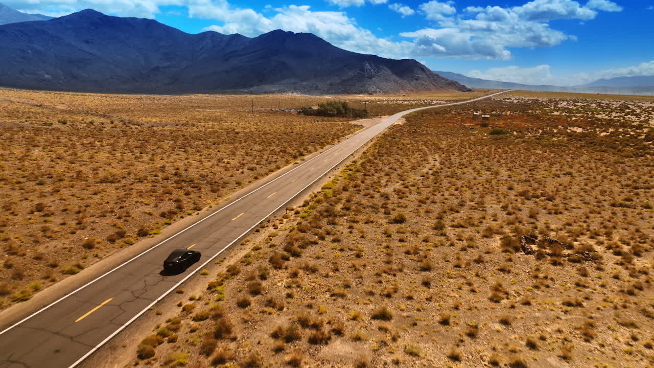 Catching up with a black car driving by the empty highway in the desert. Some huts are at the wayside. Top view.