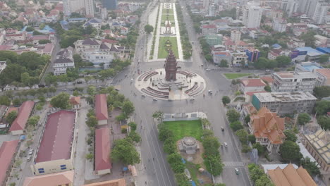 a direct overhead drone shot of phnom penh's independence monument at the heart of a traffic roundabout, flanked by historic structures and manicured greenery in the center of the boulevard