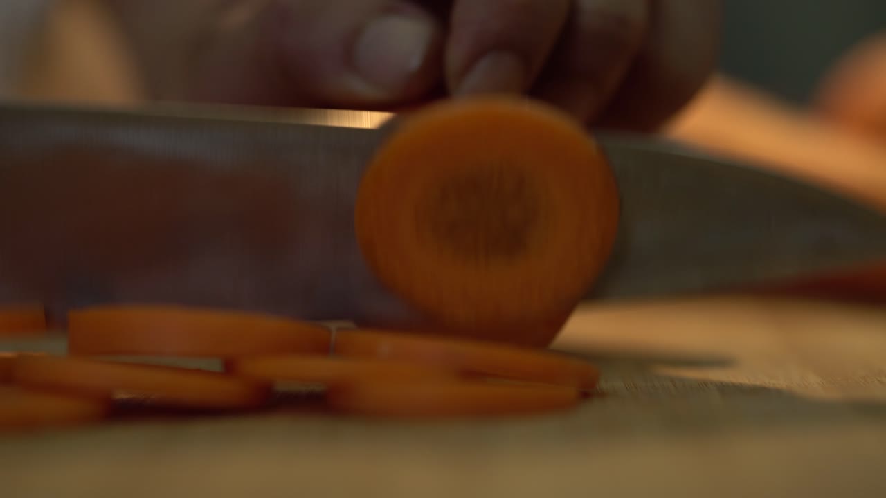 un primer plano extremo de las manos del chef cortando verduras de zanahoria peladas en rebanadas usando un cuchillo de cocina y una tabla de cortar de madera, 4k
