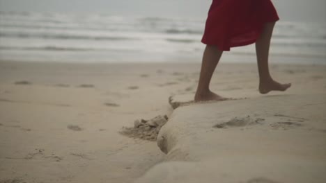 Zeitlupenaufnahme-Einer-Jungen-Frau-In-Einem-Roten-Kleid,-Die-Am-Strand-Entlang-Spaziert-Und-Mit-Ihren-Nackten-Füßen-Das-Sandige-Ufer-Streichelt