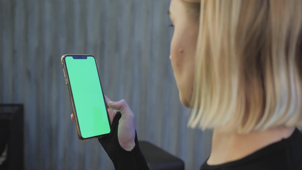 una joven atlética sostiene un teléfono inteligente en la mano, muestra una pantalla chromakey verde, está en el gimnasio