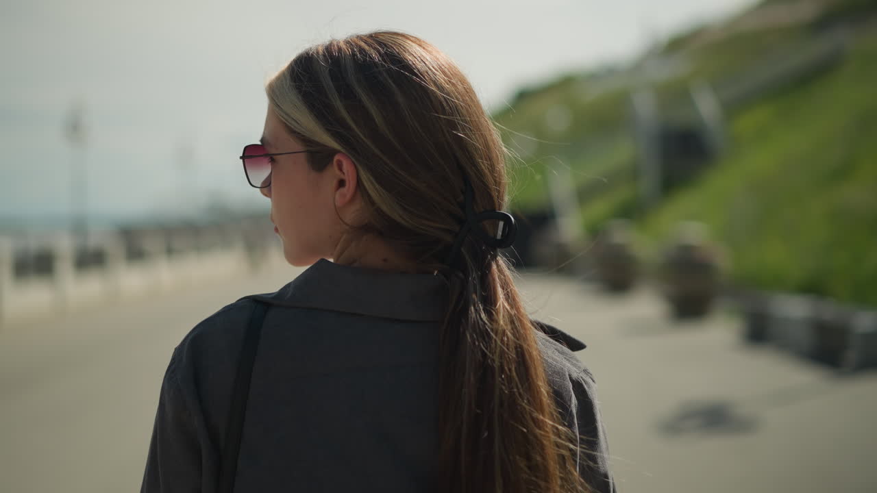 Back view of a woman walking on a tarred road with her hair tied back, looking to the left in the distance, the background is slightly blurred, with greenery and road elements visible