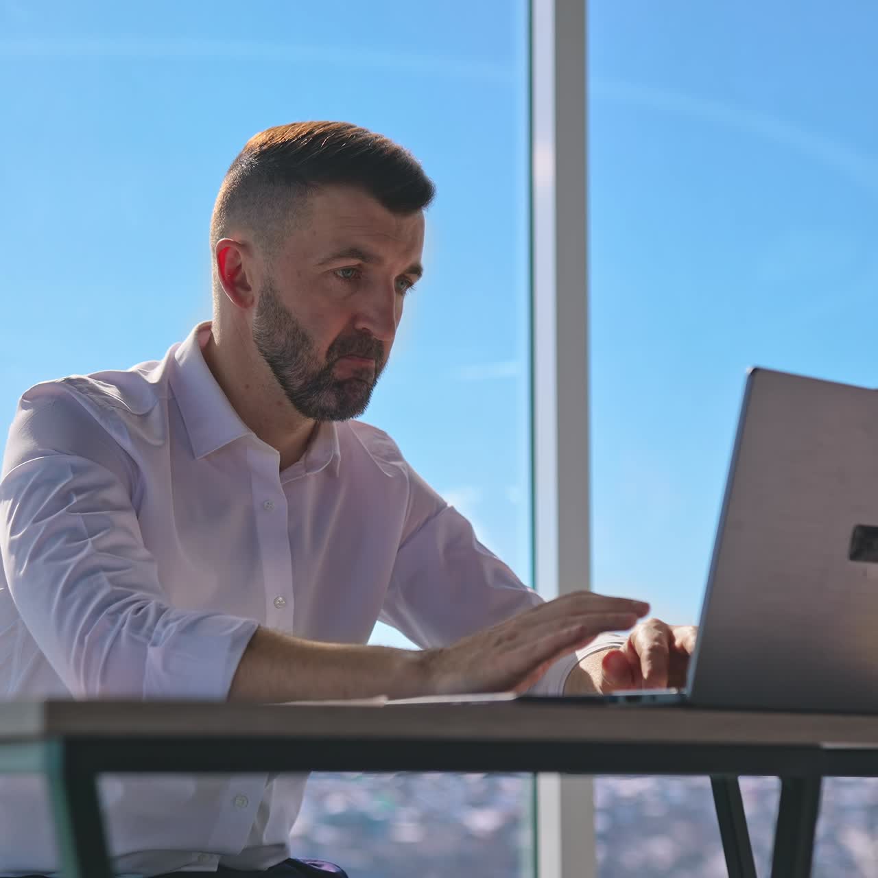 Focused businessman working on laptop computer in office with large windows. Bearded male worker using laptop computer at workplace. Handsome man looking at laptop screen in office