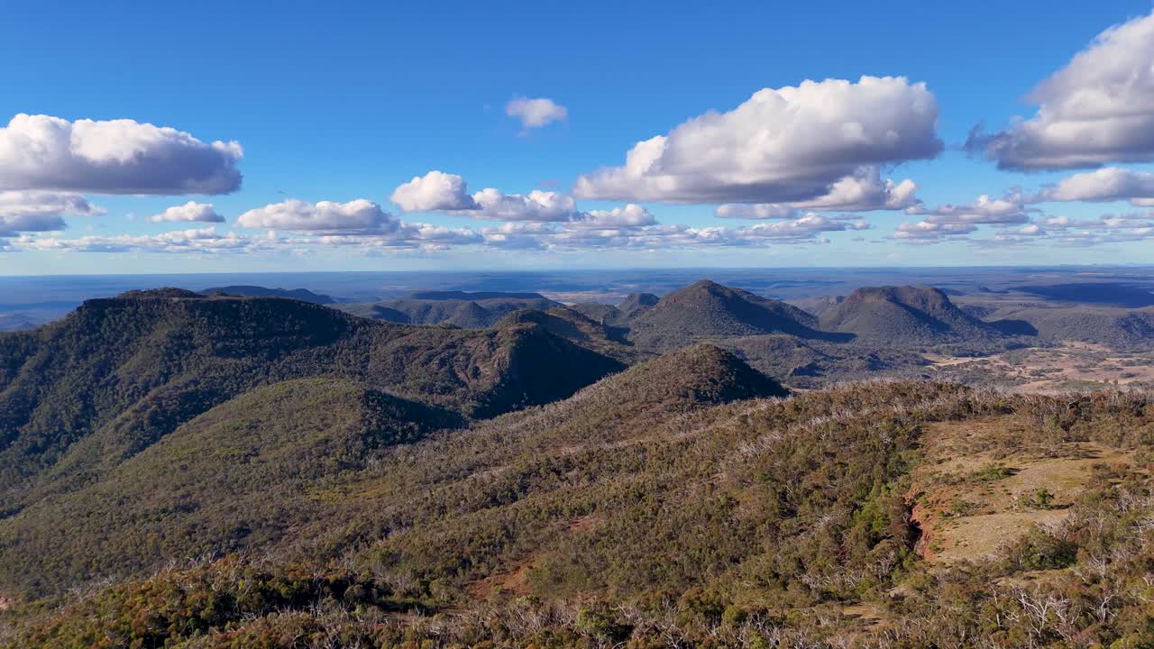 Drone camera smoothly pans over rugged mountain ridges and forested valleys in Warrumbungle National Park, under bright daylight and scattered clouds