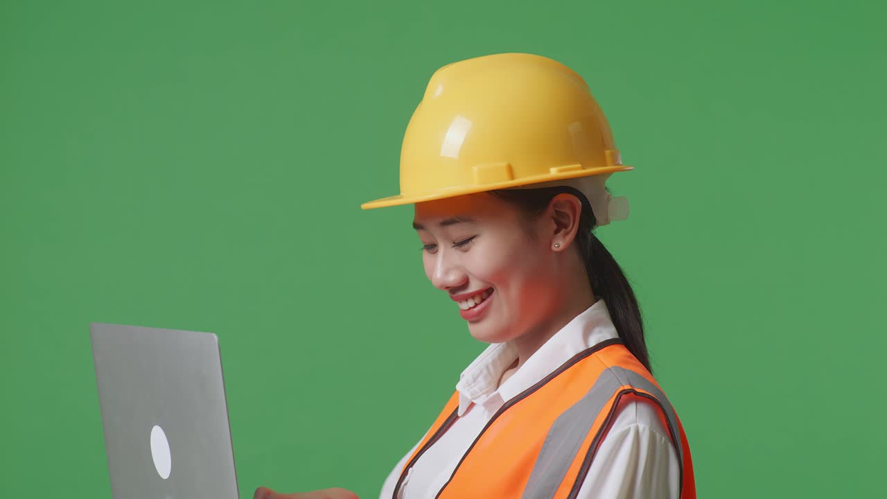 녹색 화면 배경 스튜디오에 서서 노트북에서 작업하는 안전 헬을 착용한 아시아 여성 엔지니어의 근접한 측면 시각
