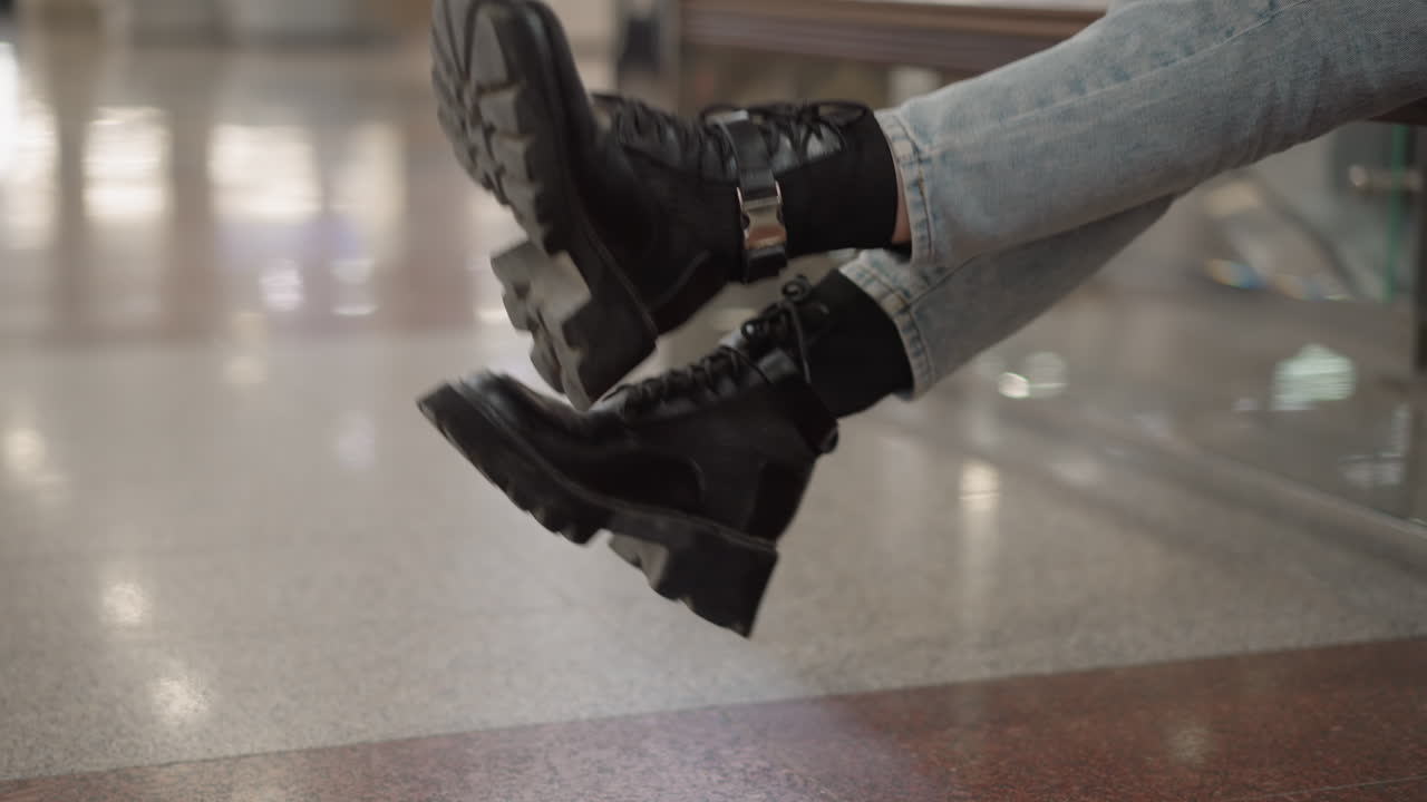 leg view of demure woman seated on mall bench, legs crossed wearing chunky black boots gently swinging against tile floor and glass railing, denim jeans visible under lighting motion