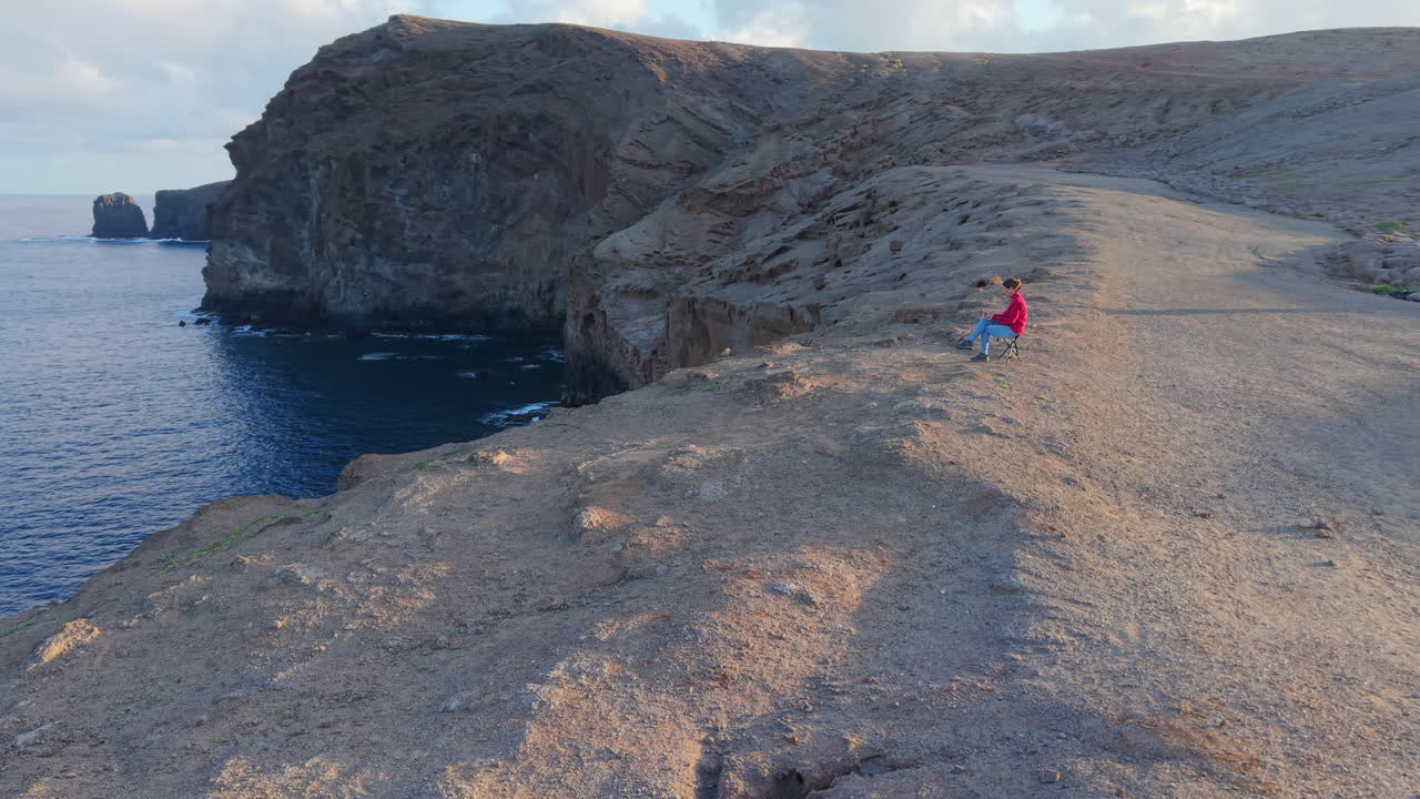 An aerial view of a woman working remotely on her computer in front of a cliff on the island of Gran Canaria. Roque Partido.
