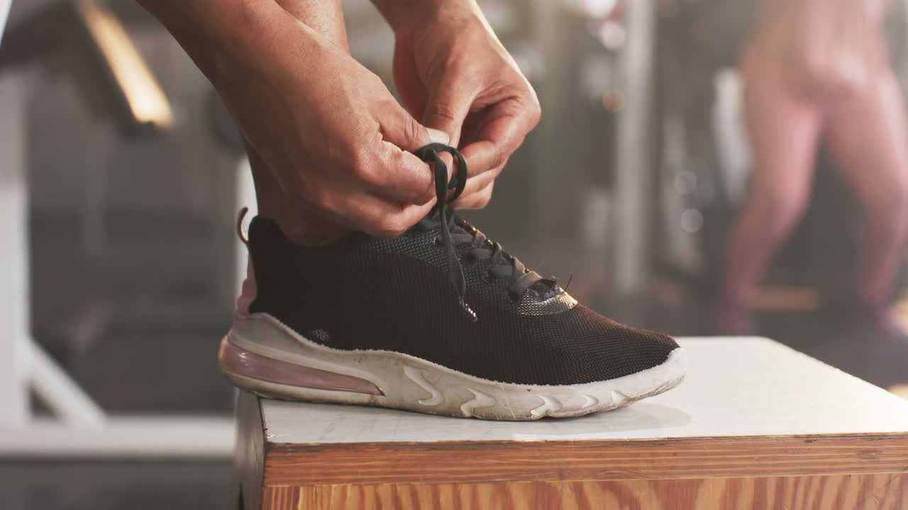 Donna afroamericana di mezza età che infila e allaccia i lacci delle scarpe nere su una pedana pliometrica in palestra durante il riscaldamento