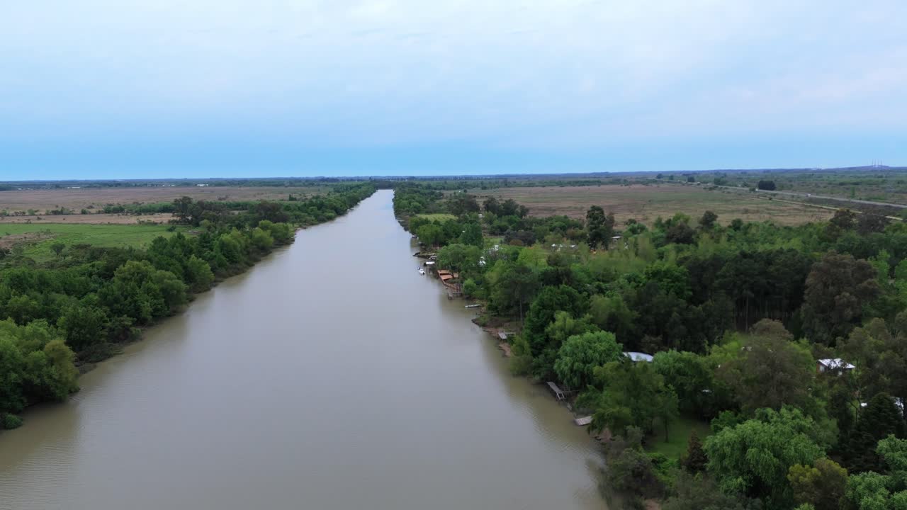 A wide aerial shot flying over the calm, brown waters of the Martín Irigoyen Canal in the Buenos Aires Delta, Argentina, on a cloudy day