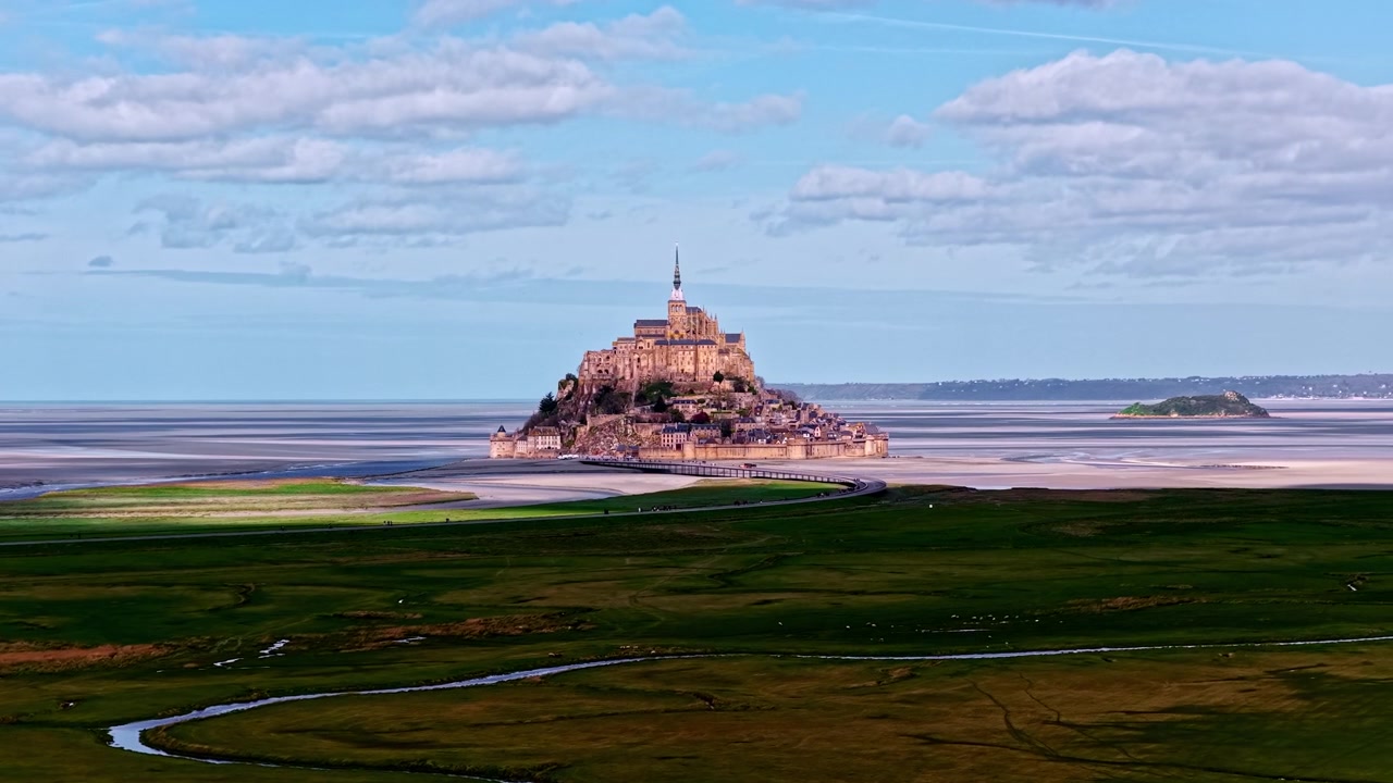 Det ikoniske Mont Saint-Michel rejser sig majestætisk mod en baggrund af frodige grønne marker og udstrakte vadehavsområder. Velegnet til rejsedokumentarer eller historiske indslag.