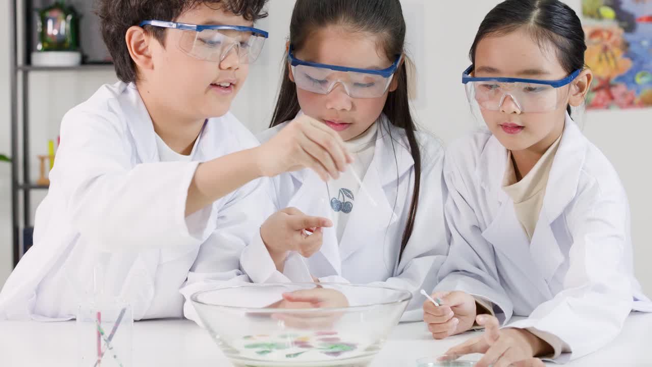 白衣を着た3人の若いアジア系の学生が、学校でカラフルな化学反応実験を行っている。