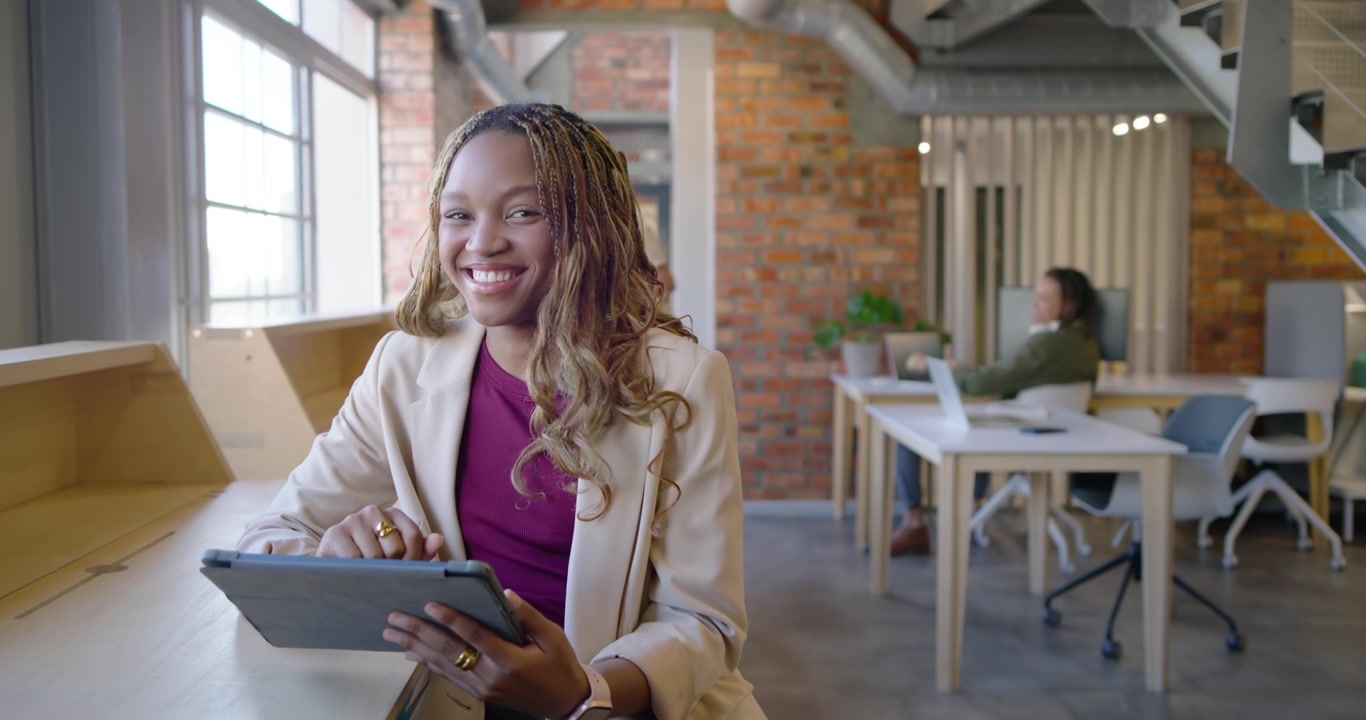 Mid-adult woman holding tablet stylus, working at window desk, noticing colleague moving, smiling