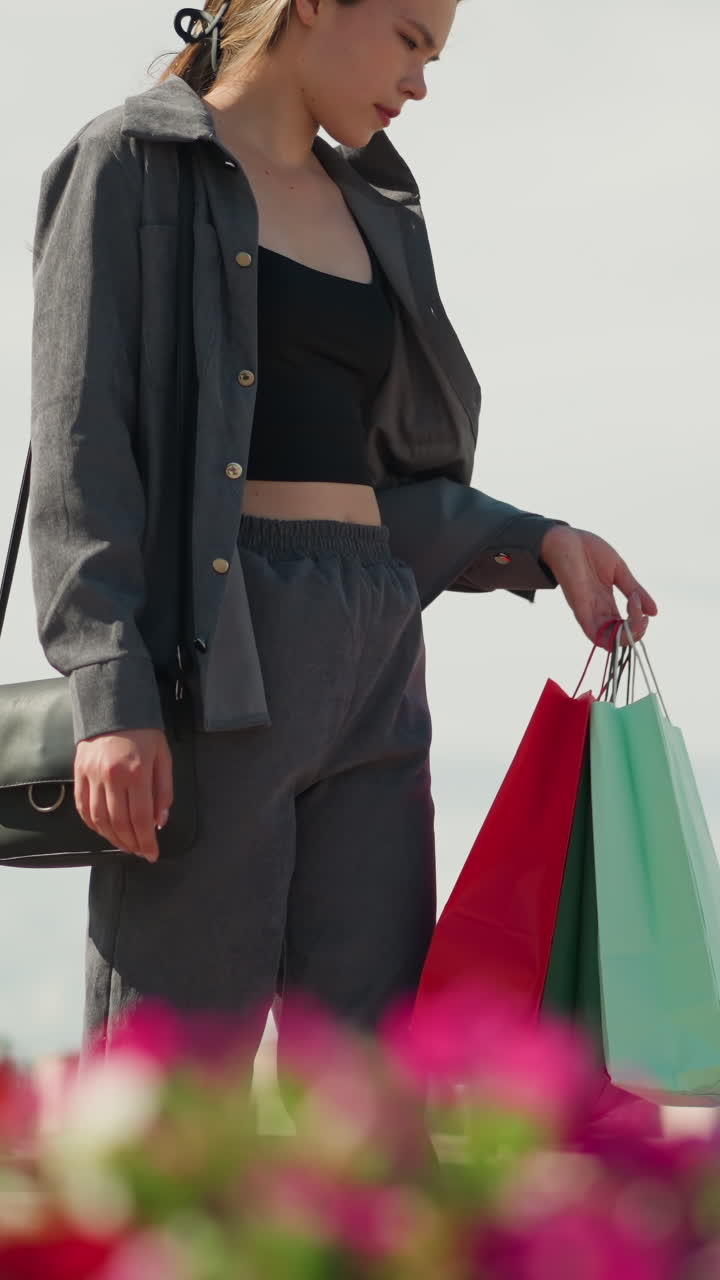 한 여인이 야외 벤치에 앉아서 초록색과 빨간색 쇼핑 가방을 에 놓고 검은색 드백을 들고 가방을 조정합니다.