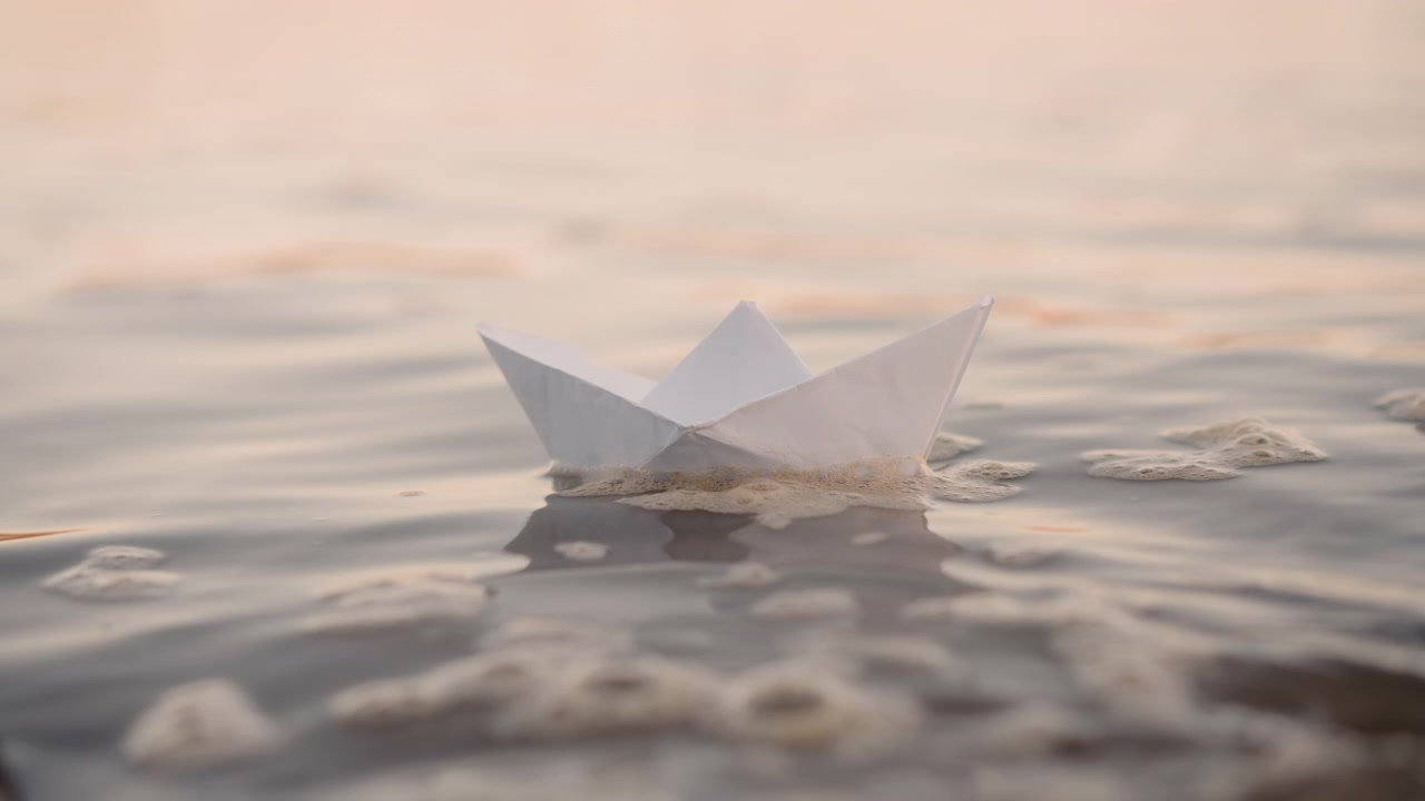 Una barchetta di carta bianca galleggia dolcemente su acqua calma, alla deriva gradualmente con leggere increspature. L'illuminazione soffusa crea un caldo riflesso sullo sfondo, enfatizzando i dettagli della texture e il movimento naturale.