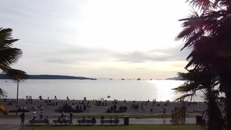 Physical-distancing-beach-aerial-pan-back-thru-palm-trees,-from-a-beach-thats-somewhat-respectable-number-of-people-clearly-distancing-each-other,-as-the-sun-sets-at-English-Bay,-BC,-Canada