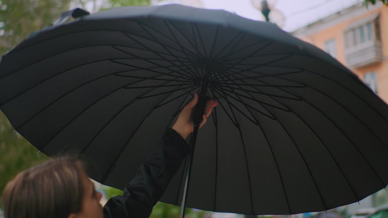 Beautiful girl in black coat as she is closing umbrella from underneath view on city street during cloudy weather with blurred background of trees and buildings showing lifestyle moment protection