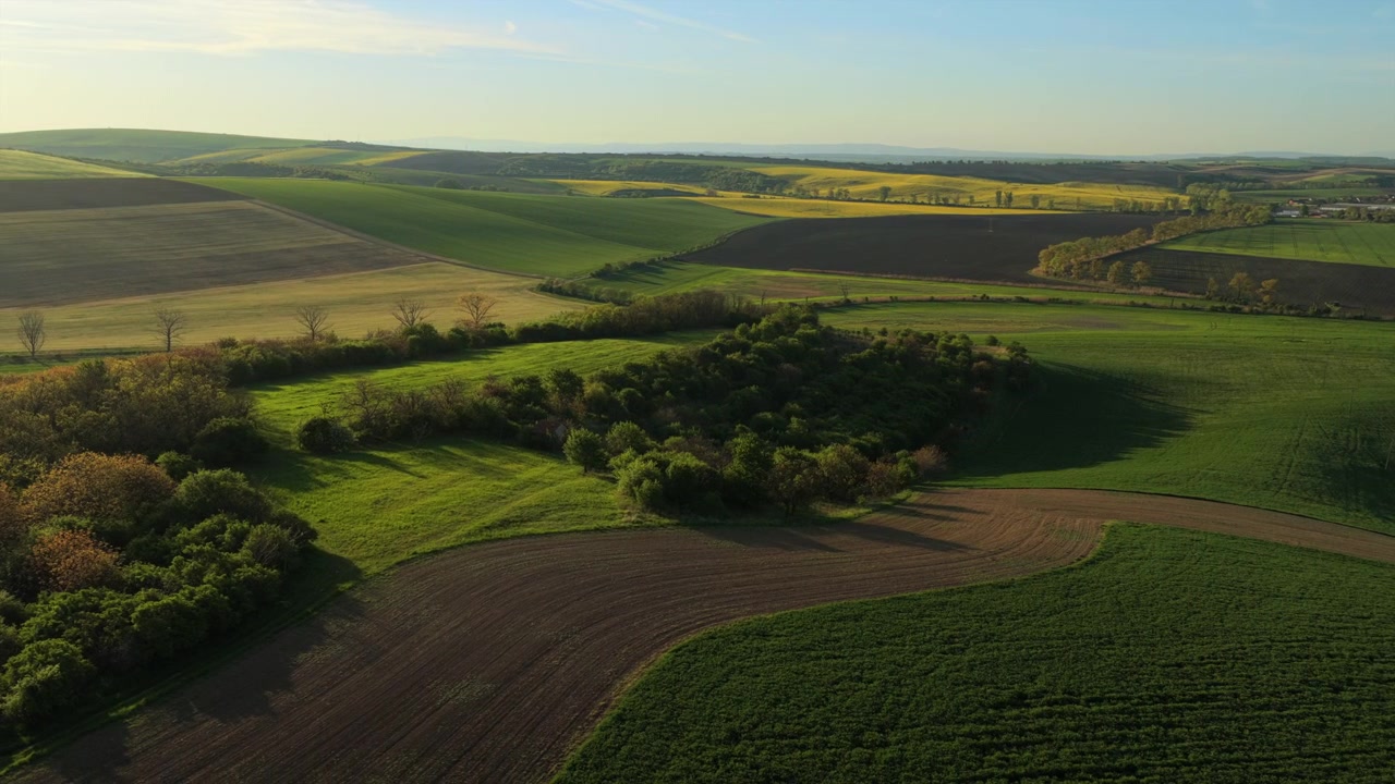 Aerial drone view of rolling Moravian farmland at golden hour, with patchwork green crop fields, plowed brown earth and scattered trees stretching to distant hills, Czech Republic.