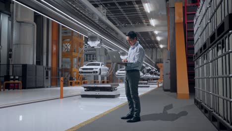 full body side view of an asian male professional worker standing with his laptop in ev car factory, high-tech industrial facility, typing on his laptop's keybaord with meditation