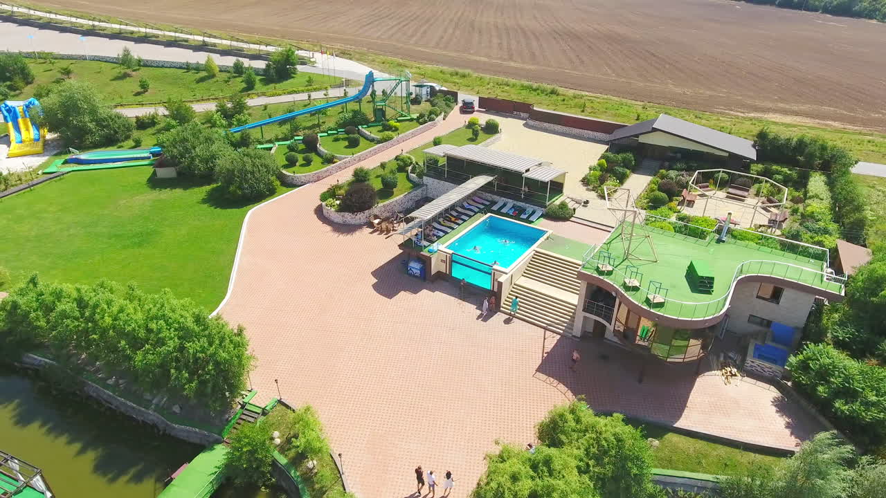 People swimming in the pool and resting around it. Aerial footage over the green beautiful resort territory. Farmlands at backdrop.