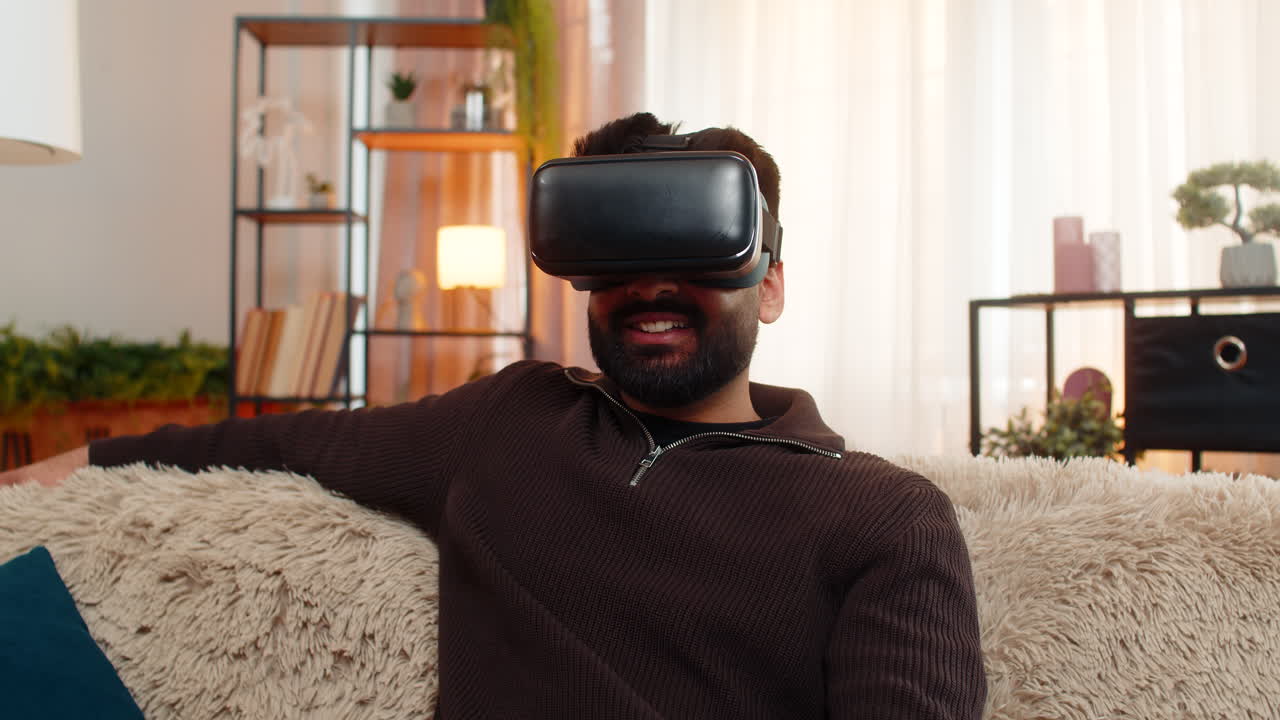 Indian man on sofa wears vr headset at home and reacts with excitement to immersive virtual world
