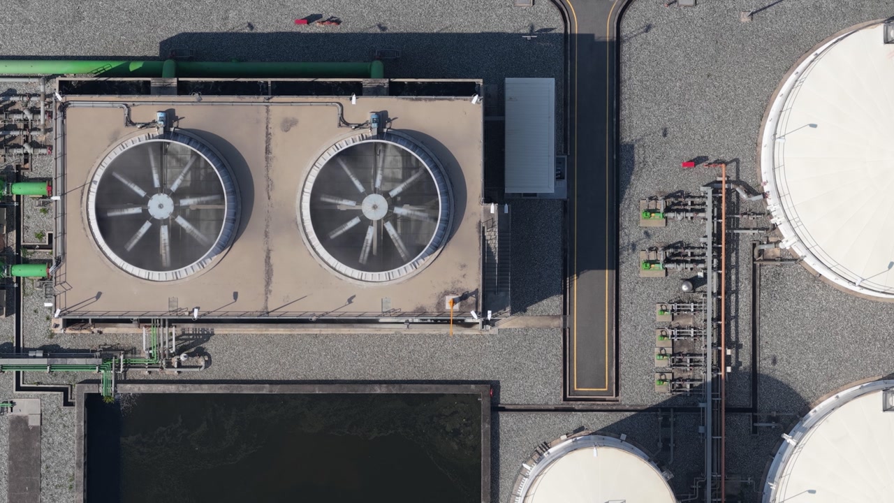 Top down aerial view of a modern industrial facility featuring large cooling towers with spinning fans, storage tanks, and a complex network of pipes, showcasing energy production