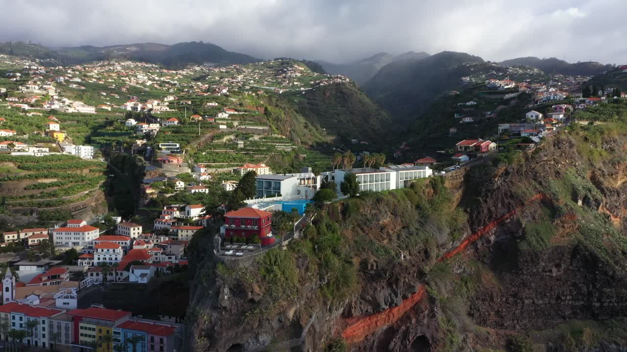 toma aérea circular de villas de lujo y un hotel en la cima de un acantilado en madeira con un pueblo en el fondo y nubes oscuras en la cima de la montaña
