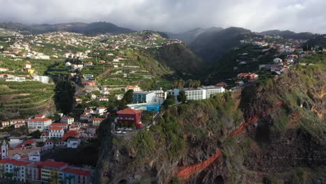 circling aerial shot of luxury villas and a hotel on top of a cliff in madeira with a village in the background and dark clouds on top of the mountain