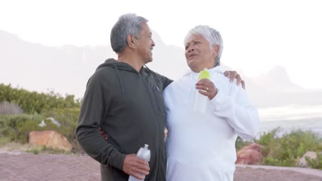 portrait of happy senior biracial couple embracing and holding bottles of water in mountains