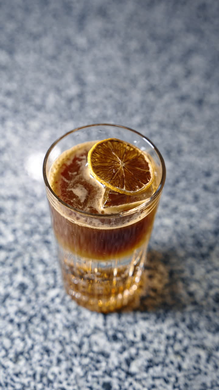 Ice cube in the glass with brown beverage. Long metal tongs putting the slice of dried orange on top of the drink. Vertical video.
