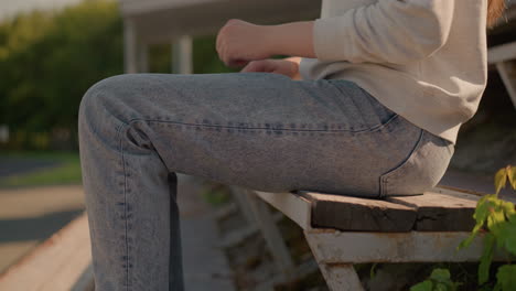 close-up of woman in jeans sitting on first row of rustic stadium seating, adjusting left leg forward, with hand resting on leg, sunlight and small green plants