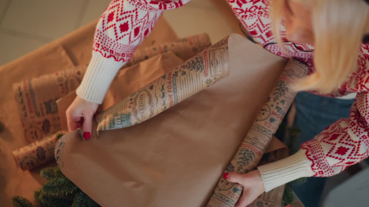 White woman wrapping christmas present with kraft paper, smoothing seams and rolling patterned sheet over package on cozy table Nordic sweater, warm lighting, scissors, tape and small greenery nearby