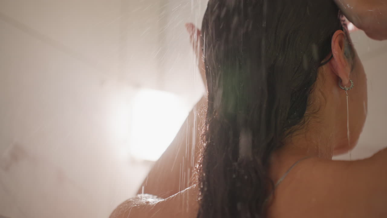 Primo piano del bagno dell'hotel con ospite che usa il dispenser di shampoo, mani che raccolgono la schiuma, flaconi e dispenser a parete nell'inquadratura, capelli bagnati adagiati sulla schiena, enfasi sui servizi dell'hotel, piastrelle moderne