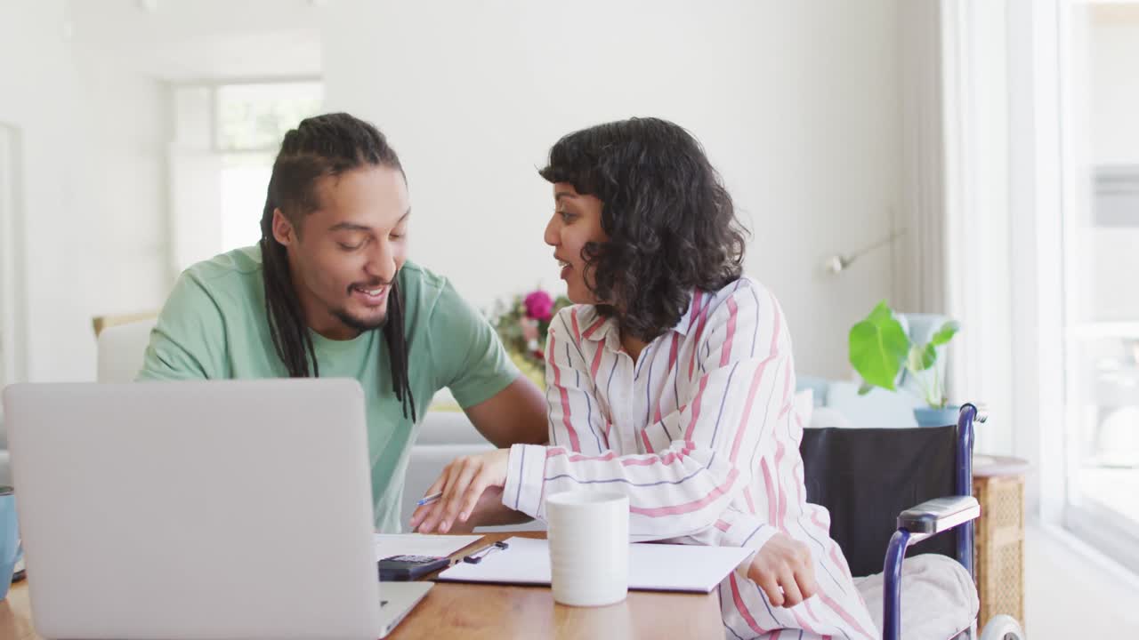 mujer biracial feliz en silla de ruedas y compañero masculino sonriente usando computadora portátil y hablando en la sala de estar