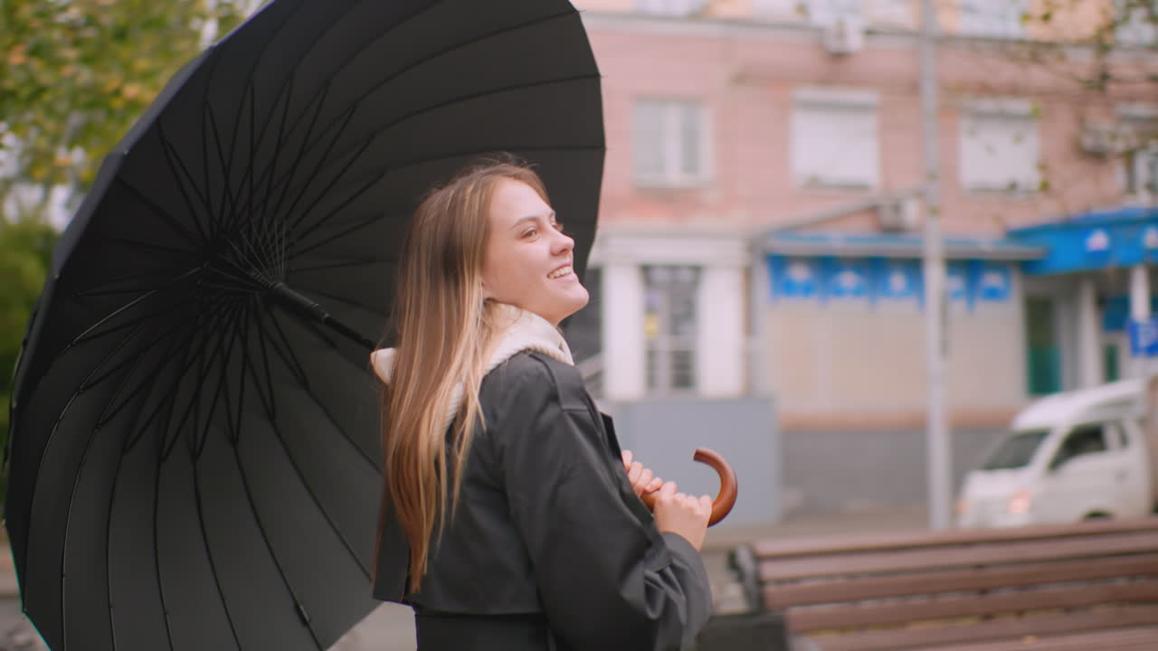 Smiling woman holding umbrella walks along city street on rainy day wearing black coat and white hoodie enjoying autumn weather with falling leaves trees buildings and cars creating urban lifestyle mood
