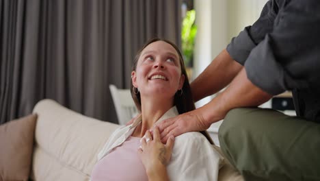 Happy-brunette-woman-sitting-on-the-sofa-while-her-husband-holds-her-shoulders-and-massages-her-at-home-on-the-weekend