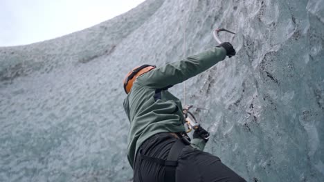 a climber in an orange helmet and specialized gear skillfully ascends a textured, blue-white ice wall with ice axes, showcasing the challenging and exhilarating sport of ice climbing in iceland