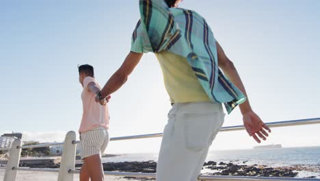 Happy-diverse-gay-male-couple-walking-and-holding-hands-at-promenade-by-the-sea,-slow-motion