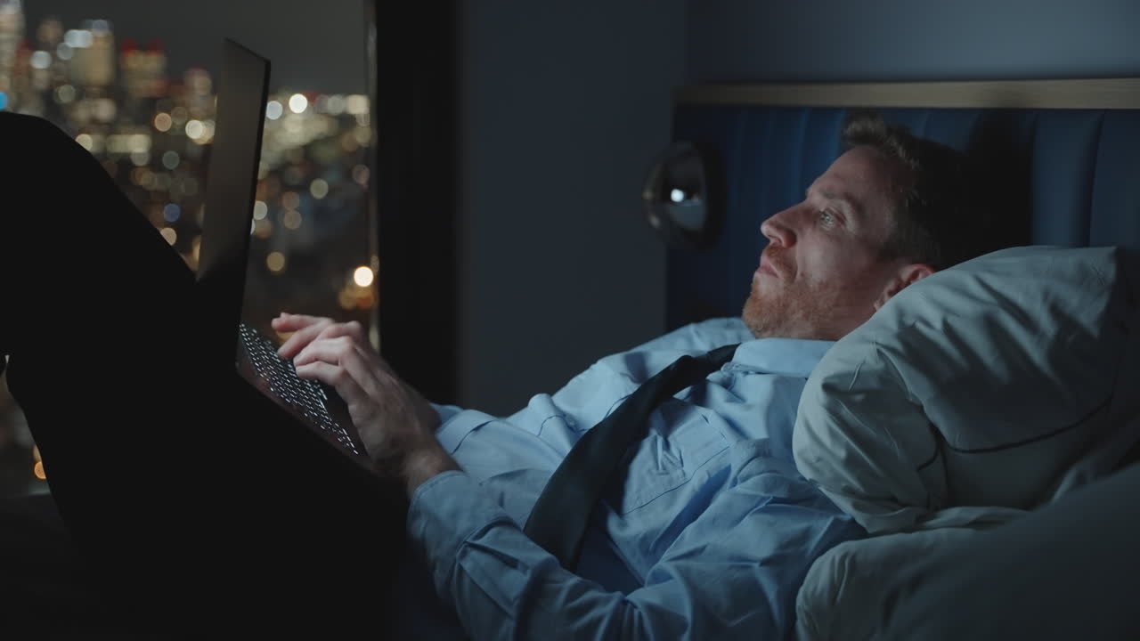 Young businessman or executive is working late into the night typing on computer laptop while in bed overlooking the city