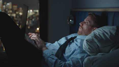 young businessman or executive is working late into the night typing on computer laptop while in bed overlooking the city