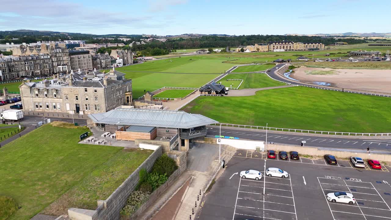Drone footage glides over a Victorian brick building, parking lot, and lush green golf course at St Andrews, Scotland, under natural daylight