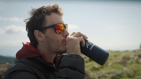 primer plano de un excursionista con lentes de sol rojos tomando un sorbo de su termo en la cima de una montaña