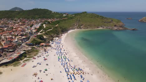 Stadt-Und-Die-Küste-Des-Meeres-Mit-Hügeln-Und-Felsen-Mit-Mehreren-Menschen,-Die-Einen-Sonnigen-Tag-Am-Strand-Mit-Sonnenschirmen-Genießen,-Ein-Wunderschöner-Ort-Arraial-Do-Cabo-Brasilien