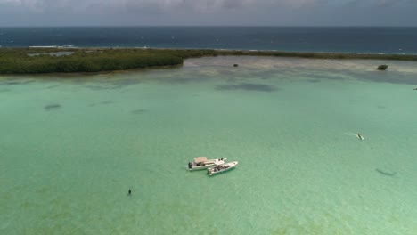 Two-boats-MOORED-waiting-kitesurfers-flying-around-turquoise-sea-waters,-LOS-ROQUES