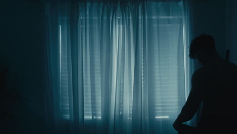 silhouette of muscular man curly hair putting shirt on in front of window curtains