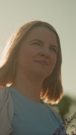 primer plano de una mujer joven sonriendo suavemente mientras sostiene un ramo de flores púrpuras, de pie al aire libre en un brillante día soleado con el sol reflejado en su cara, disfrutando de un momento de paz